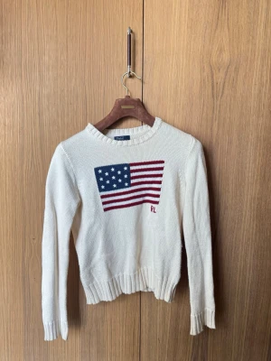Vit stickad tröja Polo Ralph Lauren - Vit stickad tröja från Polo Ralph Lauren med amerikansk flagga i blått, rött och vitt på bröstet samt RL-broderi. Tröjan har rund hals, långa ärmar och ribbade muddar. Klassisk och tidlös design i mjukt material.