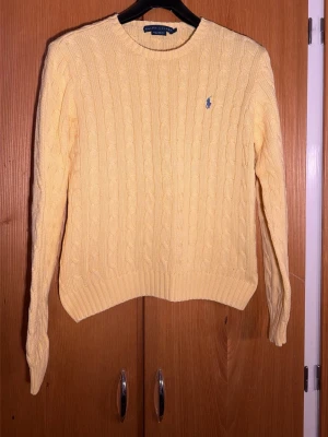Ralph Lauren Gul kabelstickad tröja  - Säljer en ljusgul kabelstickad tröja från Ralph Lauren i 100% bomull. Klassisk rund halsringning, långärmad och med den ikoniska blå broderade loggan på bröstet. Perfekt för dig som vill ha en preppy och fräsch look.