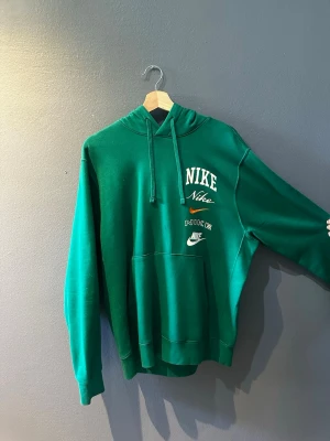 Grön hoodie från Nike med tryck - Snygg grön hoodie från Nike med flera vita logotyper tryckta på bröstet. Klassisk känguruficka framtill och justerbar huva med dragsko. Perfekt för en avslappnad och sportig stil.