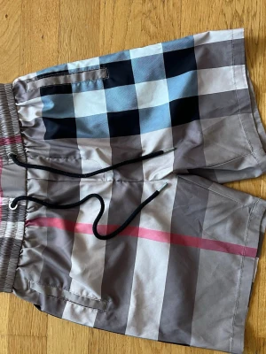 Burberry shorts - Snygga shorts med klassiskt rutigt mönster i beige, svart, blått och rosa. Shortsen har elastisk midja med svart snörning och fickor både fram och bak. Perfekta för sommardagar när du vill ha en chill och trendig look.