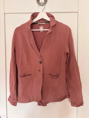 Rosa kavaj med två fickor - Snygg rosa kavaj med två framfickor och en knapp framtill. Kavajen har en avslappnad passform, mjukt material och en uppvikt krage. Perfekt för dig som vill ha en stilren men bekväm look.