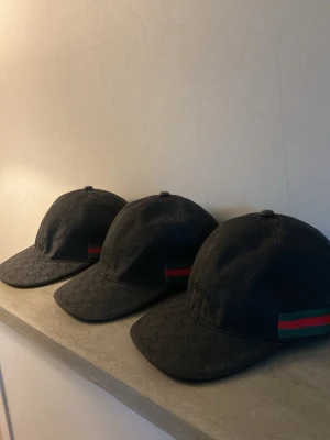 Bulk 3 stycken Svarta  kepsar  med Guc@i-logga - Snygg svart keps från Guc@i med diskret GG-monogram över hela tyget och klassiska gröna och röda ränder på sidan. Justerbar rem baktill i läder och metallspänne. Perfekt accessoar för en trendig och exklusiv look.