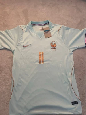 Ljusblå Frankrike fotbollströja - Snygg ljusblå fotbollströja från Nike med Frankrikes landslagsemblem och nummer 11 i guld på både fram- och baksidan. Namnet 'OLISE' står på ryggen. Tröjan har korta ärmar och tunna guldiga detaljer längs sidorna. Perfekt för match eller träning.
