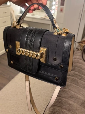 Svart handväska med gulddetaljer - Supersnygg svart handväska med gulddetaljer från river Island. Nypris 500 kr som min syster köpte ny men hjälper säljer nu vidare. Banden går att ta av och justera längden på. 💕