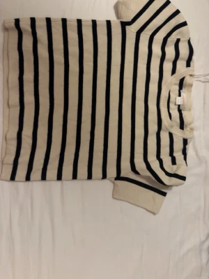 Randig topp från Gina Tricot - Kortärmad topp från Gina Tricot i vitt med mörkblå horisontella ränder. Klassisk rund halsringning och mjukt stickat material som känns skönt mot huden. Perfekt för en avslappnad och stilren look.