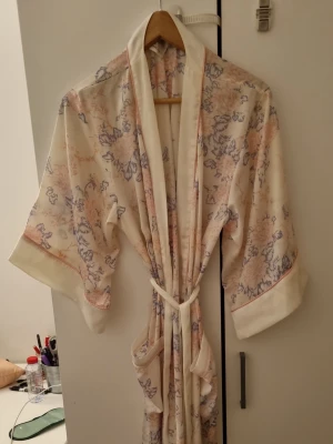 Blommig morgonrock från H&M M/L - Ljus morgonrock från H&M i storlek M/L med blommigt mönster i pastellfärger som rosa och lila. Morgonrocken har breda ärmar, vit kant och knytskärp i midjan. Tillverkad i mjukt, lätt material som känns skönt mot huden.