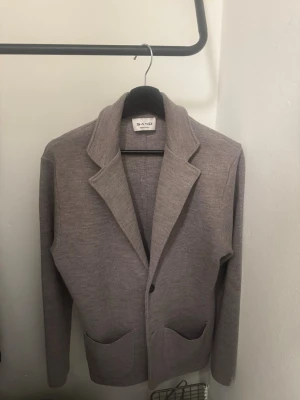 Grå kavaj från Sand - Stilren grå kavaj från Sand med två framfickor och klassisk krage. Tillverkad i ett mjukt, stickat material som ger en avslappnad men ändå dressad look. Perfekt för dig som vill ha en modern och bekväm kavaj.