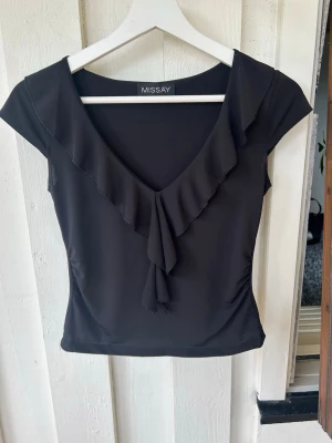 Svart topp med volang från Missay - Snygg svart topp från Missay med v-ringning och volangdetaljer längs ringningen. Toppen är ärmlös och har en figurnära passform som ger en stilren look. Perfekt att matcha med både jeans och kjol för en trendig outfit.