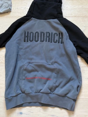 Grå och svart hoodie från Hoodrich - Säljer en grå och svart hoodie från Hoodrich med stor logga på bröstet och ficka framtill. Hoodien har huva och raglanärmar i svart, medan kroppen är grå. Praktisk magficka med dragkedja och röd textdetalj. Perfekt för en streetwear-look.