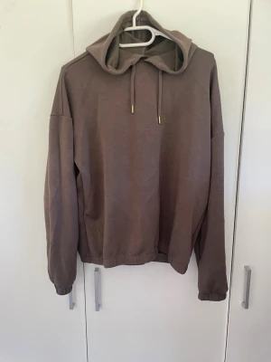 Brun hoodie med huva och dragsko - Säljer en enkel och stilren brun hoodie med huva och dragsko. Hoodien har långa ärmar, ribbade muddar och resår i nederkanten. Perfekt för chill dagar eller när du vill ha en avslappnad look. Materialet känns mjukt och bekvämt mot huden.