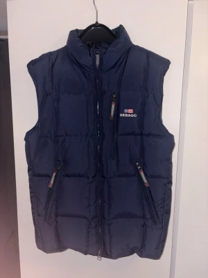 Mörkblå dunväst från Sebago - Snygg mörkblå dunväst från Sebago med hög krage och dragkedja framtill. Västen har två framfickor med dragkedja och diskret Sebago-logga med flagga på bröstet. Perfekt för lager-på-lager och håller dig varm under kyliga dagar.