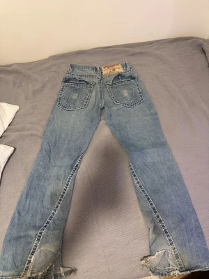 True Religion ljusblå bootcut jeans - Ljusblå jeans från True Religion med bootcut-modell och slitna detaljer vid bensluten. Stora bakfickor med den klassiska hästsko-sömmen och tydlig patch bak i midjan. Perfekta för en avslappnad streetwear-look.