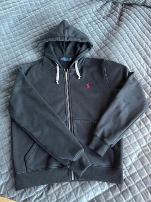 Svart hoodie från Polo Ralph Lauren - Snygg svart hoodie med dragkedja från Polo Ralph Lauren. Klassisk design med vit dragsko i huvan och röd broderad logga på bröstet. Mjuk bomullsblandning, kängurufickor och ribbade muddar vid ärmar och nederkant. Perfekt för en chill och stilren look.