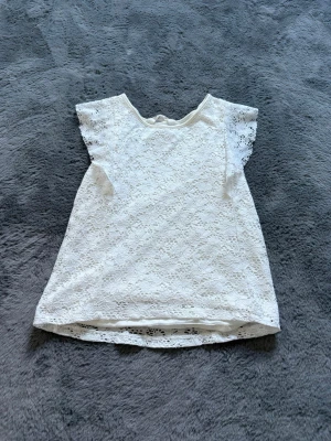 Vit spetsblus med kort ärm - Söt vit blus i spets med korta ärmar och rund hals. Blusen har ett fint spetsmönster över hela plagget och lätt volang vid ärmslut. Perfekt för dig som gillar romantisk och feminin stil.