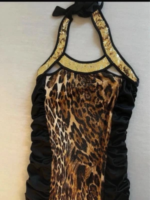 Leopardmönstrad halterneckklänning - Snygg kort halterneckklänning med leopardmönster framtill och svarta sidor. Klänningen har guldfärgade paljettdetaljer vid halsen och knyt i nacken. Perfekt för dig som vill sticka ut med ett djurmönster och glittriga detaljer.