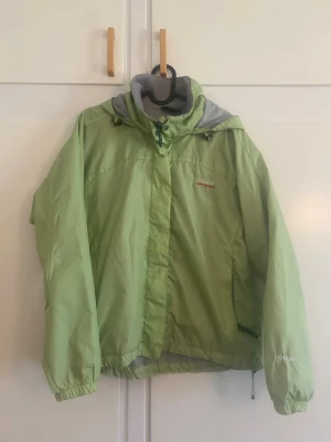 Peak Performance grön jacka, - Snygg ljusgrön vindjacka med huva och dragsko. Jackan har dragkedja framtill, elastiska muddar och gråa detaljer vid huva och insida. Märket HWent syns på ärmen och Peak på bröstet. Perfekt för blåsiga dagar och enkel att matcha.