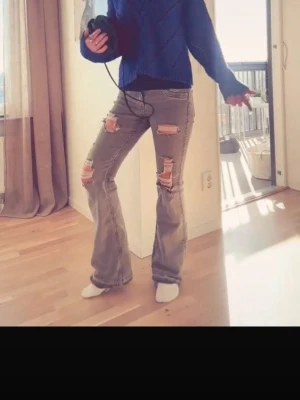 Grå slitna bootcut jeans - Säljer ett par grå bootcut jeans med stora slitningar och hål på låren och knäna. Jag är ca 176cm lång.  Jeansen har en klassisk femficksmodell och är tillverkade i denim med en lätt tvättad look. Perfekta för dig som gillar en edgy och avslappnad stil.