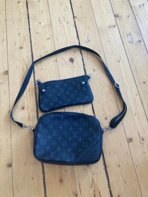 Snygg svart axelväska monogram - Snygg svart axelväska med klassiskt monogrammönster i grått. Väskan har justerbar axelrem och kommer med en matchande pouch. Tillverkad i tåligt canvasmaterial med detaljer i läder. Perfekt för dig som gillar stilrena accessoarer.