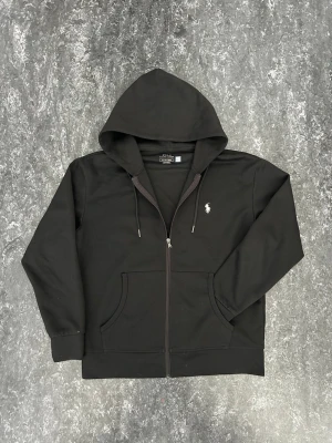 Svart hoodie från Polo Ralph Lauren - Snygg svart hoodie från Polo Ralph Lauren med dragkedja, huva med dragsko och klassisk vit logga på bröstet. Tillverkad i mjuk bomullsblandning och har två fickor framtill. Perfekt för en chill och stilren look.