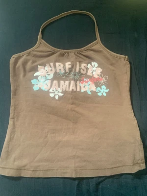 Brun linnetopp med blommigt tryck - Snygg brun linnetopp med tunna axelband och blommigt tryck framtill där det står 'Surf Isle Jamaica'. Toppen har en avslappnad passform och är perfekt för varma dagar. Materialet känns mjukt och bekvämt mot huden.