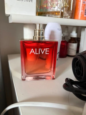 ALIVE parfym röd glasflaska - Säljer en ALIVE parfym i en snygg fyrkantig glasflaska med röd till orange toning och guldfärgad spraykork. Flaskan har en modern och stilren design som gör sig snygg på hyllan. Perfekt för dig som vill ha en fräsch och elegant doft. Used only a few times and personally I’m not into the smell