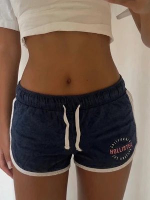 Blå shorts med snörning från Hollister - Mörkblå shorts från Hollister med vita detaljer och snörning i midjan. Shortsen har vit kant runt benen och logga på vänster sida. Perfekta för sommaren och riktigt sköna att bära till vardags eller träning.