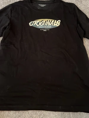 Svart t-shirt från Jack & Jones - Snygg svart t-shirt från Jack & Jones med trycket 'ORIGINALS' i vitt och beige på bröstet. Klassisk passform och rund halsringning, perfekt till jeans eller shorts. Tillverkad i mjuk bomull för skön känsla hela dagen.