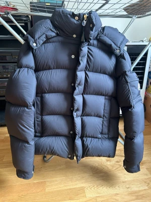 Svart puffig dunjacka med huva - Säljer en svart dunjacka med stor puffig passform och avtagbar huva. Jackan har hög krage, dragkedja och knappar framtill samt en ficka på ärmen. Perfekt för kalla vinterdagar och riktigt varm tack vare tjockt dunfoder.