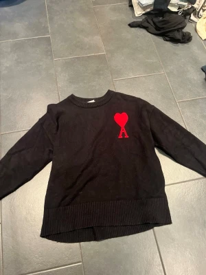Ami Paris tröja  - Säljer en svart stickad tröja med rund hals och ribbade muddar. På bröstet finns ett rött hjärta ovanför bokstaven A, vilket ger en unik och trendig look. Perfekt för dig som gillar stilrena plagg med en twist.
