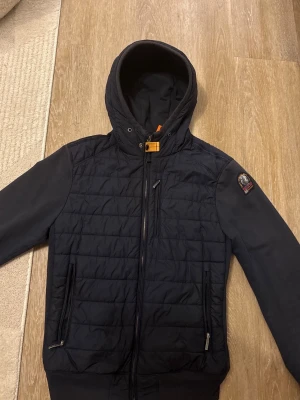 Parajumper’s vindjacka  - Snygg mörblå vindjacka från Parajumpers med quiltad front, hel dragkedja och två fickor med dragkedja. Jackan har en fast huva, ribbade muddar och en patch på ärmen. Insidan har ljusgrått foder och praktiska innerfickor med orange detaljer.
