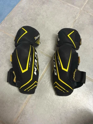 CCM Tacks armbågsskydd svart/gul - Säljer ett par svarta och gula armbågsskydd från CCM Tacks-serien. Skydden har justerbara kardborreband och slitstark textilyta med gula detaljer. 🌸