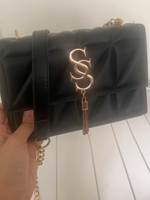 Svart quiltad axelväska med gulddetaljer - Snygg svart axelväska i quiltat mönster med guldfärgad kedjerem och dekorativt SSS-emblem framtill. Väskan har en elegant tofsdetalj och stilren design som passar till många outfits. Perfekt för dig som gillar trendiga accessoarer.