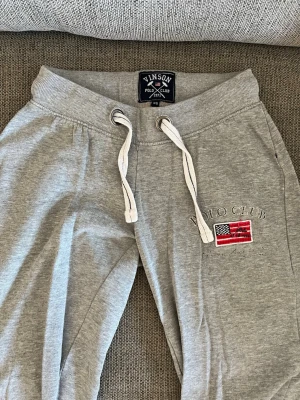 Grå mjukisbyxor Vinson Polo Club XS - Grå mjukisbyxor från Vinson Polo Club med broderad amerikansk flagga och text på vänster ben. Byxorna har vita dragsnören i midjan och bred mudd. Perfekta för chill dagar och har en klassisk, sportig look.