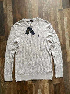 Ljusgrå kabelstickad tröja - Snygg ljusgrå kabelstickad tröja med rund halsringning och diskret broderad logga på bröstet. Tröjan har långa ärmar och ribbade muddar vid ärmslut och nederkant. Perfekt för en stilren och klassisk look.