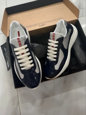  sneakers i blått och vitt - Snygga sneakers i blankt mörkblått och vitt med grå mesh-detaljer. Klassisk låg modell med vita snören och tydlig röd logga på plösen. Skorna har en sportig och modern look med kontrasterande materialval.