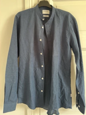 Blå linneskjorta Lindbergh - Säljer en stilren blå skjorta från Lindbergh i slim fit-modell. Skjortan är tillverkad i en mix av bomull och linne, har långa ärmar, ståkrage och vita knappar framtill. Perfekt för dig som gillar en enkel men snygg look. (Pris: 399kr)