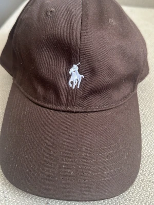 Brun keps från Polo Ralph Lauren - Snygg brun keps från Polo Ralph Lauren med klassisk böjd skärm och justerbart spänne bak. Kepsen har den ikoniska vita broderade loggan med polospelare framtill. Tillverkad i bomullstyg som känns både slitstarkt och bekvämt.