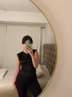 Svart ärmlös satinlinnetopp - Snygg svart ärmlös topp i satin med rund hals och figurnära passform. Perfekt att styla med både jeans och kjol för en trendig look. Materialet ger en lyxig känsla och lätt glans.