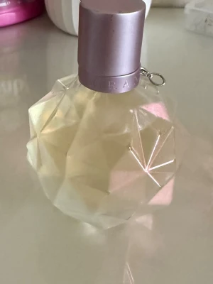 Ariana grande parfym - Säljer en parfym i en unik, facettslipad glasflaska med ljusgul vätska och ett matt silverfärgat lock. Flaskan har en modern, geometrisk design som sticker ut på sminkbordet. Perfekt för dig som gillar snygga detaljer i badrummet.