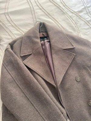Kort grå kappa med bred krage - Snygg kort kappa i grått med bred krage och dubbelknäppning framtill. Kappan har raka fickor och ett mjukt, ulligt material som ger en stilren look. Perfekt för dig som vill ha en trendig och klassisk jacka till hösten eller vintern.