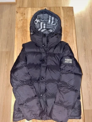 Svart dunväst med huva Burberry - Snygg svart dunväst från Burberry med avtagbara ärmar och huva. Västen har dragkedja och knappar framtill, två fickor med dragkedja och klassiskt rutigt foder i huvan. Perfekt för lager-på-lager och streetstyle.