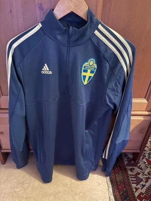 Blå Sverige träningströja Adidas - Mörkblå träningströja från Adidas med Sveriges landslagsmärke på bröstet och vita ränder längs axlarna. Halvlång dragkedja framtill och hög krage. Perfekt för fotbollsträning eller sportiga dagar.