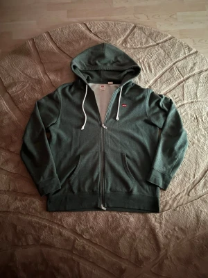Mörkgrön zip hoodie från Levi's - Säljer en mörkgrön hoodie från Levi's med dragkedja framtill och vit dragsko i huvan. Klassisk röd Levi's-logga på bröstet och två fickor framtill. Tillverkad i mjukt bomullsmaterial som känns skönt mot huden. Perfekt för en avslappnad stil.