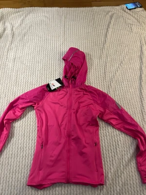 Rosa vindjacka med huva - Snygg rosa vindjacka med dragkedja framtill och huva. Jackan har två sidofickor med dragkedja och en sportig look. Perfekt för blåsiga dagar och passar till en aktiv livsstil. Materialet är tunt och smidigt, vilket gör den lätt att bära med sig. Pris lapp på