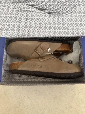 Taupe Birkenstock Boston tofflor - Snygga beige Birkenstock Boston tofflor i mocka med justerbart spänne och klassisk korksula. Perfekta för en chill och stilren look. Mjukt innermaterial och greppvänlig yttersula för extra komfort. ‼️Skickar inom 24H‼️ **PRIS GÅR ATT DISKUTERAS**
