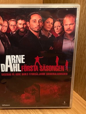 Arne Dahl Första Säsongen DVD-box - DVD-box med första säsongen av Arne Dahl, baserad på de populära kriminalromanerna. Omslaget är svart och rött med bilder på skådespelarna. Innehåller 5 filmer, totalt 900 minuter speltid. Perfekt för dig som gillar spännande thrillers och svensk krim.