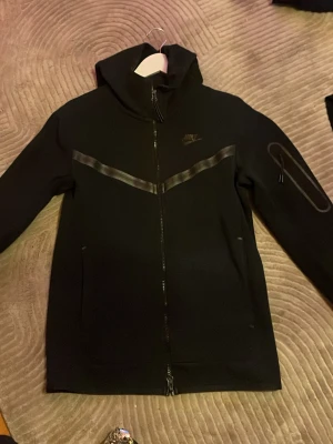 Nike Tech Fleece XS - Svart hoodie från Nike i storlek XS med hel dragkedja och huva. Snyggt mönster med reflekterande V-formad detalj över bröstet och diskret Nike-logga. Fickor på sidorna och extra detalj på ena ärmen. Perfekt för en sportig och stilren look.