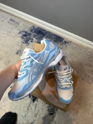 Asics gel nyc - Snygga sneakers från Asics i ljusblått och vitt med meshpaneler och syntetdetaljer. Skorna har vita snören, grå sula och klassisk Asics-logga på plösen. Perfekta för dig som gillar sportig och fräsch stil.