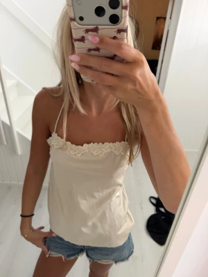 Beige linnetopp med spetsdetalj - Säljer en söt beige linnetopp med tunna axelband och spetsdetaljer längs urringningen. Toppen har öppen rygg och ett mjukt, följsamt material som känns perfekt för sommaren. Passar till både jeans och kjol för en chill look.