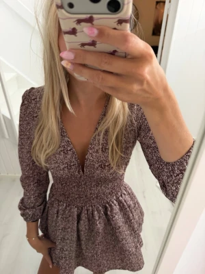 Kort blommig klänning med v-ringning - Söt kort klänning med småblommigt mönster i vinrött och vitt. Klänningen har v-ringad hals, långärmade ärmar och smockad midja som ger en fin siluett. Perfekt för dig som gillar en feminin och trendig stil.